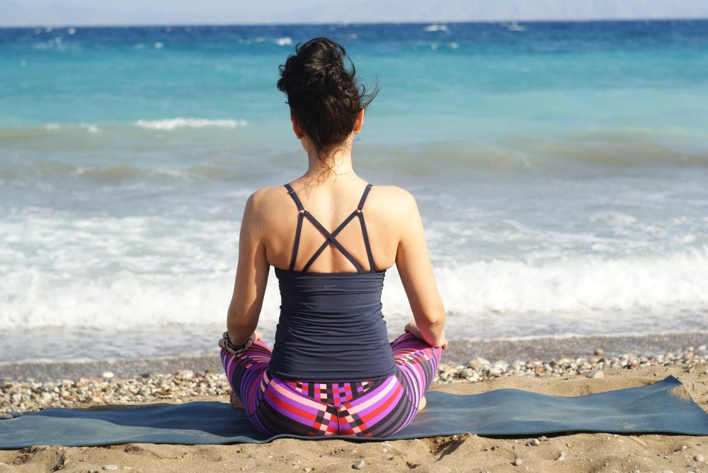 Eine Frau in Sportkleidung sitzt auf dem Strand am Meer im Schneidersitz auf einer Matte