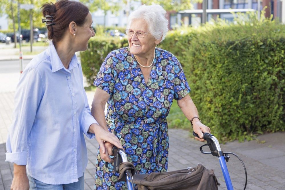 Eine lächelnde Seniorin draußen mit einem Rollator unterhält sich mit einer jüngeren Frau