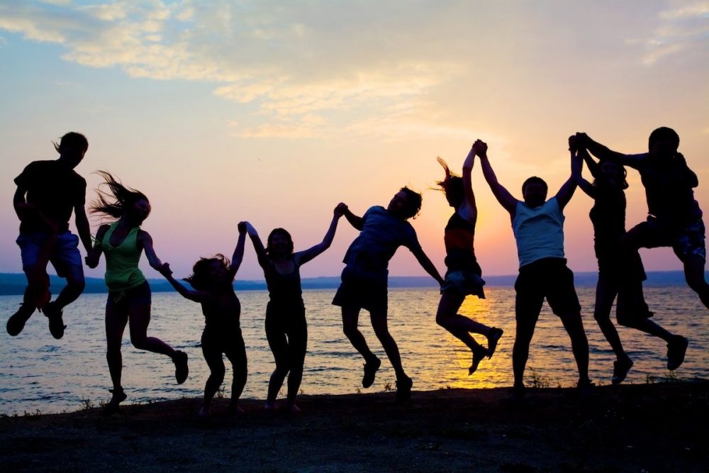 Sihlouetten einer Gruppe von Hände haltenden Menschen die beim Sonnenuntergang hochspringen